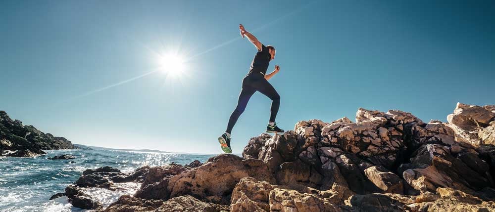 A person leaps across coastal rocks under a bright, clear sky, capturing a sense of adventure and a commitment to a healthy lifestyle