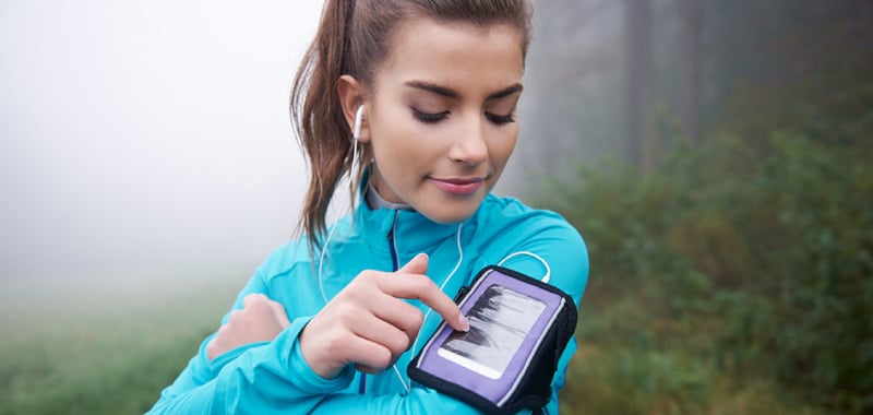 Una mujer vestida con ropa deportiva usa un teléfono inteligente atado a su brazo durante una carrera al aire libre para monitorear su salud y rendimiento físico