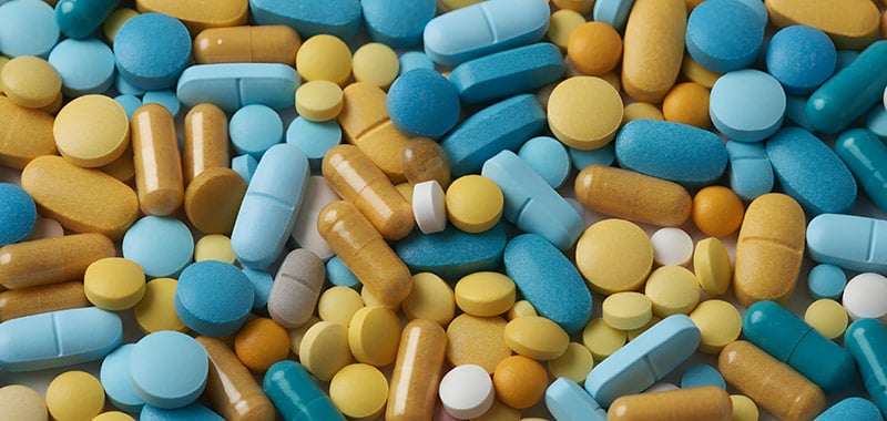 Una toma área que llena el encuadre de una pila densa de diversas píldoras, cápsulas y tabletas de suplementos en tonos de azul, amarillo y blanco.