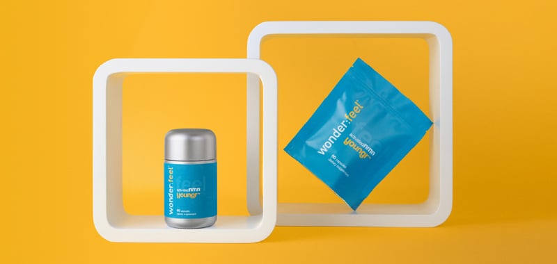 Un frasco plateado y una bolsa azul de suplementos Wonderfeel Youngr™ NMN exhibidos dentro de marcos cuadrados blancos sobre un fondo amarillo.