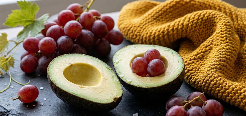 Un aguacate partido por la mitad y racimos de uvas rojas sobre una superficie de piedra oscura junto a una tela de punto amarilla texturizada, que representan alimentos integrales saludables y nutrición para la longevidad