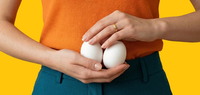 A close-up of a person's hands gently cradling and protecting two white eggs against their lower abdomen. The person is wearing an orange top and teal trousers against a vibrant yellow background. The image serves as a symbolic representation of fertility, ovarian health, or menopause.