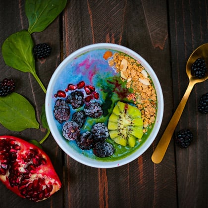 Vista superior de un colorido bol de smoothie en un recipiente blanco sobre una mesa de madera oscura. El bol está decorado con moras, semillas de granada, rodajas de kiwi y granola sobre una base vibrante de color azul y verde. Se aprecian una granada cortada y hojas de espinaca frescas alrededor.