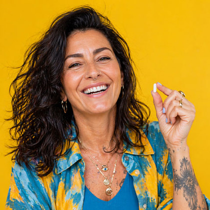 Retrato de una mujer sonriente de mediana edad con cabello oscuro y rizado sobre un fondo amarillo brillante. Ella sostiene una pequeña cápsula blanca de suplemento entre sus dedos, representando la suplementación nutricional y el envejecimiento proactivo.