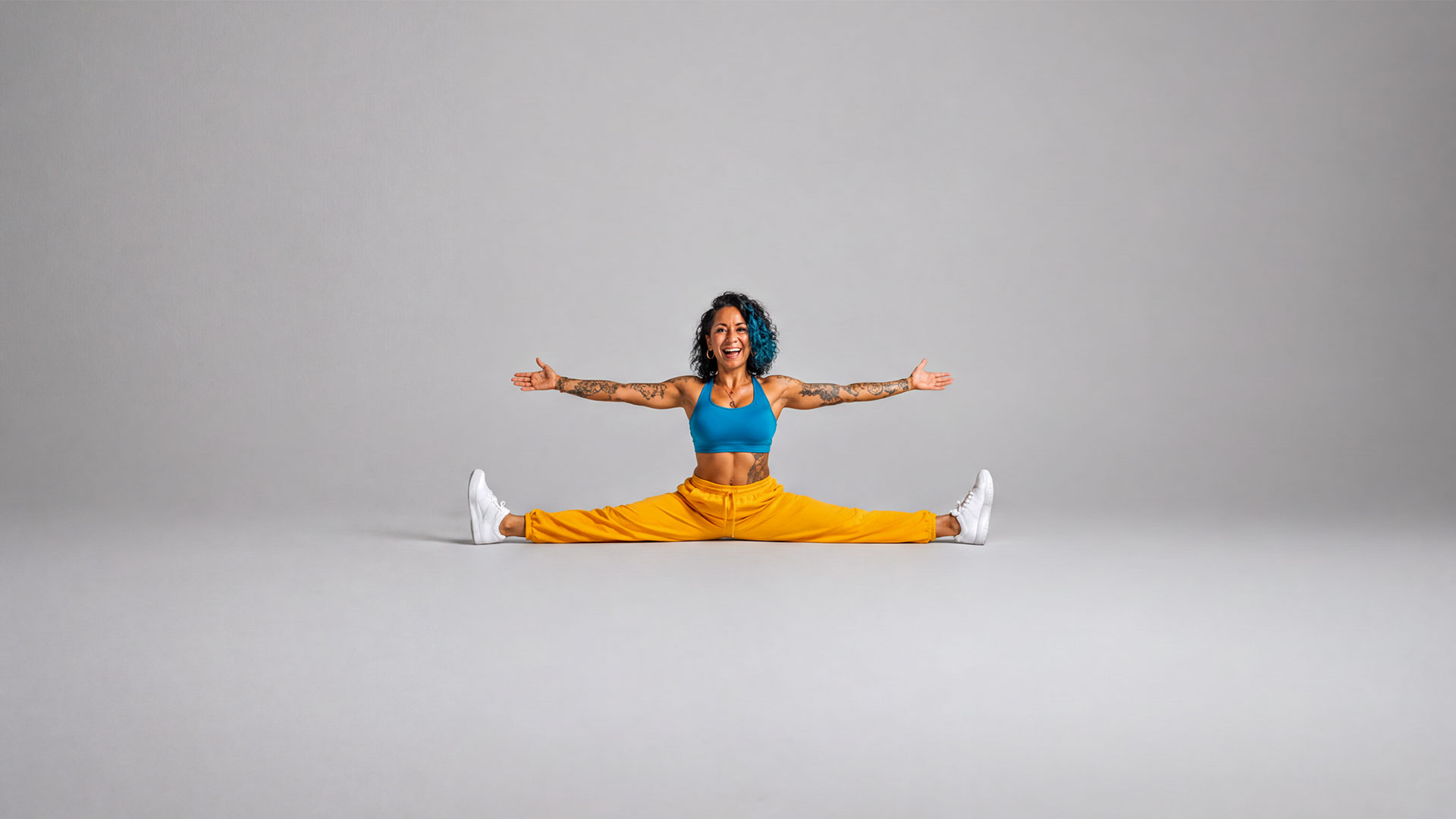 Tattooed woman with blue curly hair doing the splits with arms outstretched and a big smile, wearing a blue sports top and yellow pants against a gray background.