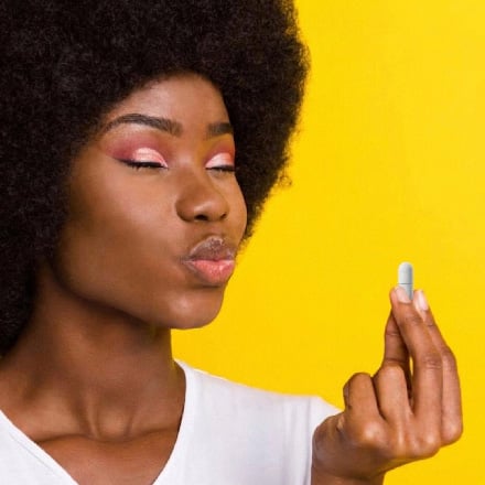 Woman with natural hair holding a supplement capsule against a bright yellow background, eyes closed in a playful expression.